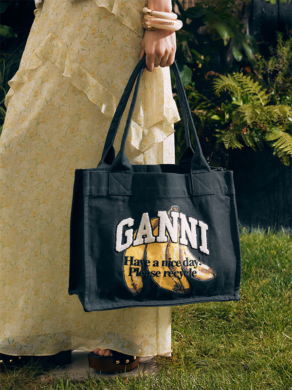 Person holding a black tote bag with 'Ganni' branding and text outdoors.
