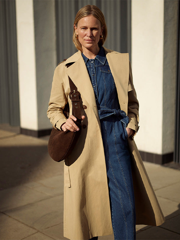 Woman wearing a beige trench coat over a blue denim outfit, holding a brown leather bag.